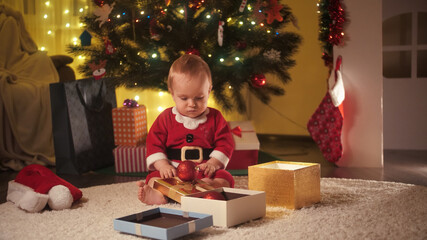Fototapeta na wymiar Happy smiling cheerful baby boy opening Christmas presents and crawling on floor under Christmas tree in living room. Families and children celebrating winter holidays.