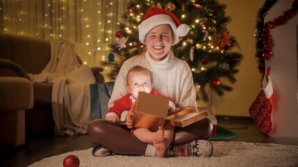 Fototapeta na wymiar Happy smiling mother holding Christmas gift and baby son while sitting on carpet under Christmas tree