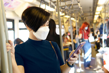 A beautiful smart look asian woman with disposable face mask on a city public train using smartphone to join her social media while travel to office. Urban, New normal, Lifestyle, Technology, Covid 19
