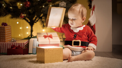 Fototapeta na wymiar Cute baby boy sitting under Christmas tree and opening lid of golden gift box