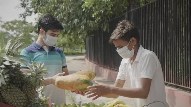 A young Indian Asian attractive looking male buying a Papaya from a local Fruit selling vendor boy on a corner of a streetside both of them wearing protective disposable mask on the face amid Covid 19