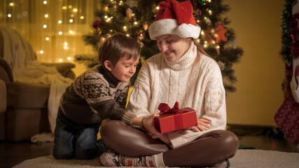 Fototapeta na wymiar Cheerful smiling boy with mother giving Christmas presents and hugging under Christmas tree at house. Families and children celebrating winter holidays.