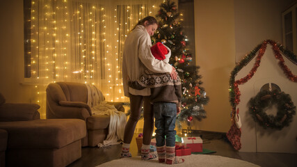 Fototapeta na wymiar Mother with son celebrating Christmas looking on glowing Christmas tree in living room. Pure emotions of families and children celebrating winter holidays.