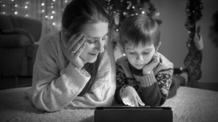 Black and white portrait of boy with mom lying on floor and using tablet computer next to Christmas tree at living room.