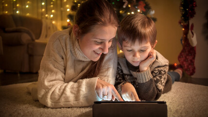 Portrait of smiling mom with son browsing internet and watching video on tablet under Christmas tree. Pure emotions of families and children celebrating winter holidays.