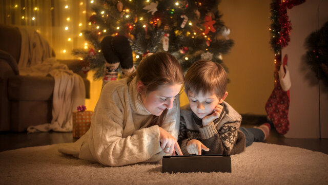 Portrait of boy with mother warm wool sweater lying on floor and using digital tablet computer on Christmas eve at house