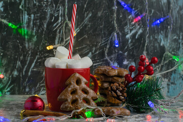 Fototapeta na wymiar Aromatic hot winter cocoa drink with chewing marshmallows in a red cup with a straw, delicious gingerbread cookies, christmas decor and a garland on a dark background, side view