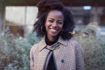 Beautiful african american young woman with afro and large hoop earrings in a stylish coat in a spring park, smiling © Bougie Banana