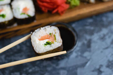 Hand holds wooden sticks sushi roll with spinach, cheese, salmon and nori. Large set of rolls on the background