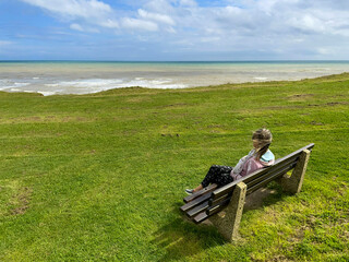 A lady sits on a wooden railed bench and concrete supports on a green grass cliff top looking out to a sea and skyline with blue sky and clouds.Wind is blowing her hair dramatically