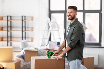 moving, people and real estate concept - happy smiling man with adhesive tape packing box at new home
