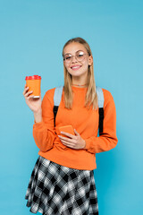 Positive student in eyeglasses holding smartphone and paper cup isolated on blue