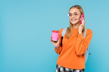 Smiling student in eyeglasses talking on cellphone and holding coffee to go isolated on blue