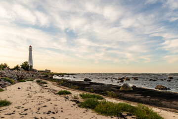 Fototapeta premium Tahkuna lighthouse on the shore during sundown at Hiiumaa, Estonia, Europe White lighthouse with red top on the rocky beach at dusk. Sun is setting near lighthouse on the coast.