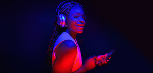  Young African American woman wearing headphones listening to music and dancing in futuristic...