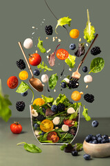 Salad with flying ingredients to prepare it on a green background. Organic salad of cherry tomatoes, baby lettuce, beets, arugula, mozzarella, blackberries, blueberries and pine nuts. 