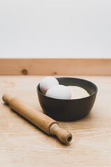 dark stoneware bowl with flour and white eggs and rolling pin on a rustic wooden tabletop