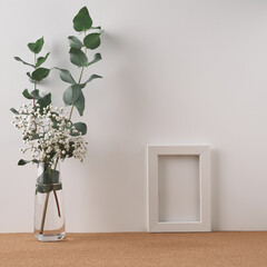 A frame and a vase with eucalyptus and white flowers stand on a cork, white background. Mock up, copy space. Folk.