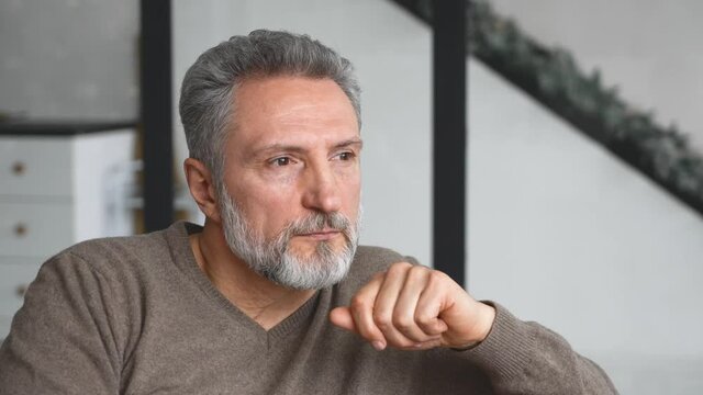 Portrait of serene and carefree handsome middle-aged grey-haired man sitting on the couch and looks aside lost in thoughts and dreams, then look at camera. Peaceful well-looking mature male resting