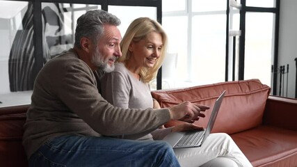 Middle-aged couple spends leisure time with a laptop at home. Spouses sit on the couch, watching video, web surfing or shopping online together, enjoying time online - Powered by Adobe