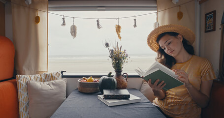 Young woman wearing hat in campervan reading book with beatiful ocean view
