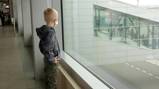 Little boy waiting boarding to flight in airport transit hall and looking through the window at airplane near departure gate.