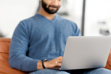 Cropped picture of Indian bearded man using laptop, Hispanic guy is typing on the keyboard, answering emails, multiracial male freelancer in casual wear working remotely from home