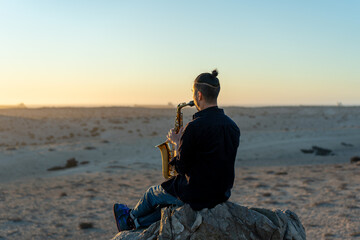 musician sitting on the rock and playing saxophone at sunset in the desert - obrazy, fototapety, plakaty