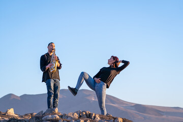 latin musician is playing saxophone while latin woman is dancing in the desert landscape at sunset	