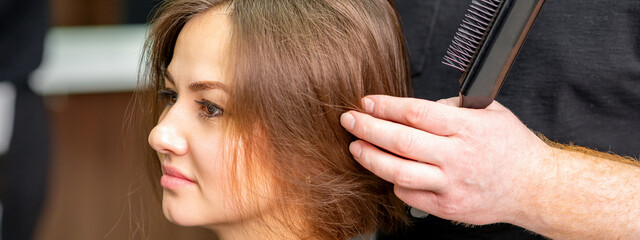 Naklejka premium Male hairdresser works on the hairstyle of the young caucasian brunette woman at a hair salon