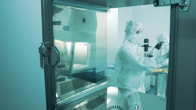 Handheld shot of two Caucasian female scientists in full protective coveralls with hood, putting a blood plasma bag and test tube rack with samples in the pass-through chamber.