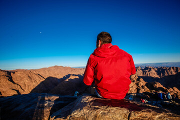 A man in a red jacket sits facing the rising sun on Mount Sinai in Egypt.