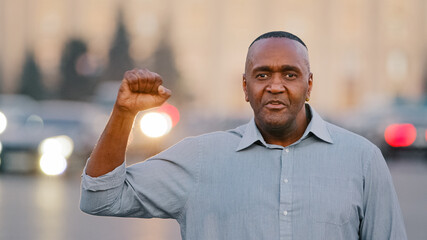 Confident Black African American retirement age man protests against discrimination standing looking at camera talking showing with hand gestures fist against racism racial discrimination solidarity