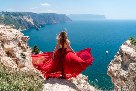 A girl with loose hair in a long red dress descends the stairs between the yellow rocks overlooking the sea. A rock can be seen in the sea. Sunny path on the sea from the rising sun