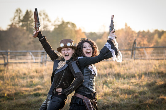 Two women in steampunk costumes with fake pistols rejoice and point gun barrels up
