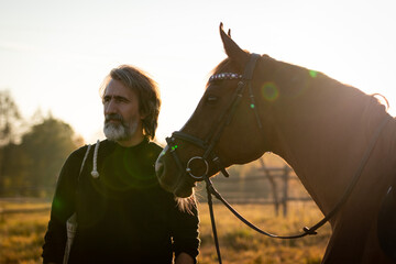 Mature man with a beard on a ranch in the fall at sunset next to a horse
