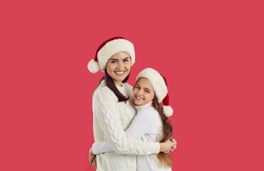 Fototapeta premium Portrait of smiling young Caucasian mother and small teen daughter in Santa hats isolate don red studio background. Happy mom and little girl child celebrate Christmas together. New Year concept.
