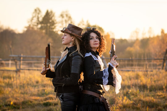 Two women in steampunk suits with fake pistols stand back to back against an autumn landscape at a ranch.