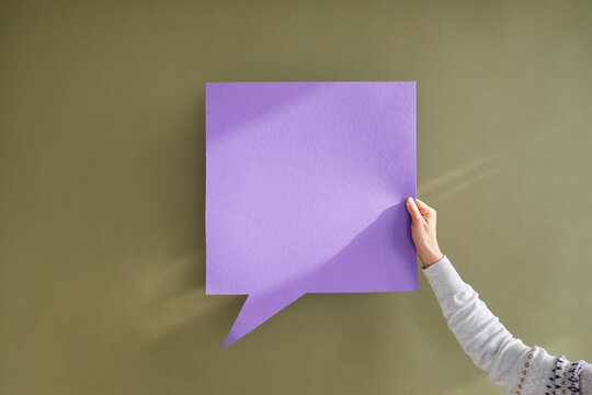 Anonymous adult showing one purple square shaped mockup copyspace speech bubble in hand on greyish green background. Free, blank, clean violet paper for expressing opinion and getting message across - Powered by Adobe