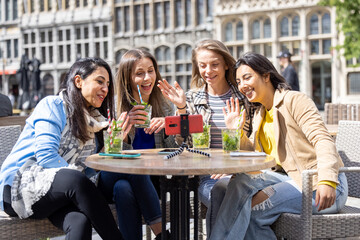 Antwerp, Belgium, May 21, 2021, four mixed race female tourists or students sitting outside in the old city center at a cafe terrace holding a video call using mobile phone. High quality photo