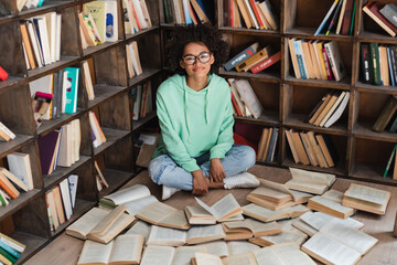 pleased african american student in eyeglasses sitting with crossed legs surrounded by books in...