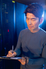 Asian male engineer writing on clipboard in computer server room