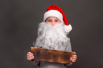 a boy 8-10 years old in a hat and with a Santa beard. He holds a wooden sign in his hands for advertising and Christmas sale. on a gray background. The concept of New Year sales. copy space