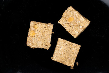 Three pieces of halva with peanuts on a black ceramic dish, close-up, top view.