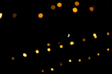 Night sky with crescent moon surrounded by orange-yellow bokeh glow from decoration string lights 
