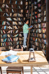 african american student in eyeglasses standing on wooden ladder while reading book near blurred...