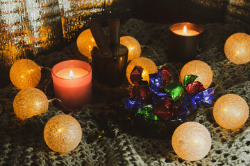 Christmas still life with sweets, two candles, a garland in the form of balls woven from threads. The still life is located on a knitted scarf, which gives the photo an atmosphere and comfort. 