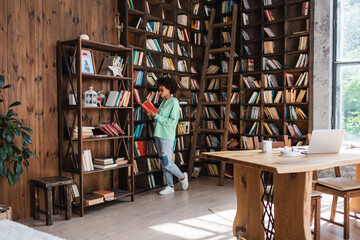 full length of young african american student in eyeglasses reading book near gadgets on desk