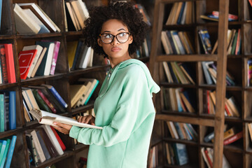 young african american student in eyeglasses holding book near bookcase