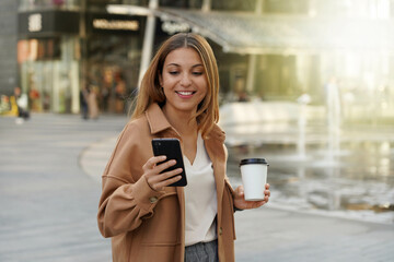 Entrepreneur young. Busy woman. Smartphone app. Office worker outdoors. Cappuccino coffee cup. Shopping mall. Architecture modern. Urban background.
